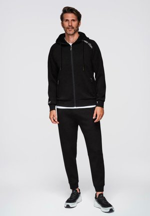 Uomo con felpa nera con cerniera, pantaloni da jogging neri, scarpe da ginnastica bianche e nere, in piedi davanti a uno sfondo grigio uniforme.