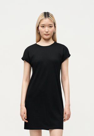 Giovane donna con capelli biondi lisci che indossa un vestito nero a maniche corte, in piedi davanti a uno sfondo chiaro e uniforme.