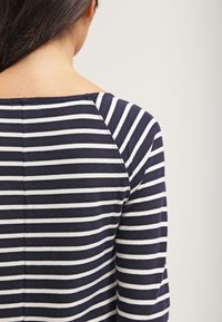 Femme aux longs cheveux foncés portant un t-shirt à rayures horizontales bleu marine et blanc, vue de dos sur un fond clair uni.