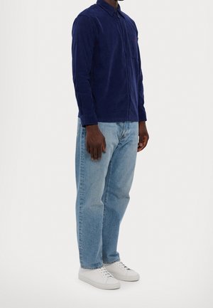 Homme portant une chemise à manches longues bleu foncé, un jean bleu clair et des baskets blanches, debout devant un fond clair uni.