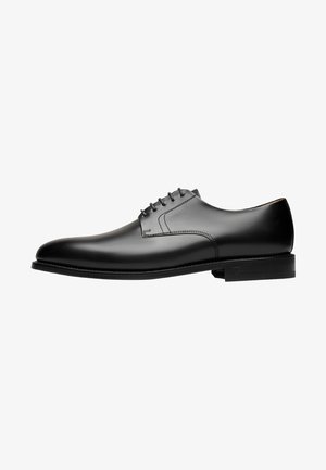 Chaussure habillée en cuir noir avec un bout arrondi, un minimum de coutures, six œillets pour les lacets et une semelle en caoutchouc plate et légèrement texturée.