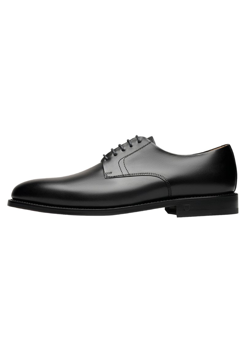Chaussure habillée en cuir noir avec un bout arrondi, un minimum de coutures, six œillets pour les lacets et une semelle en caoutchouc plate et légèrement texturée.