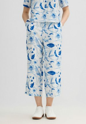 Persona que lleva pantalones blancos de pierna ancha y top a juego con estampado botánico azul, combinados con zapatos blancos sobre fondo claro.