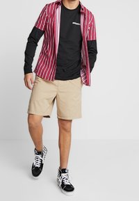 Rot-weißer gestreifter Kurzarmshirt über einem schwarzen Langarmshirt, kombiniert mit khakifarbenen Shorts und schwarz-weiß karierten Sneakern.