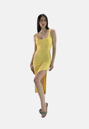 Donna in piedi, vestita con un abito giallo aderente con uno spacco alto e sandali intrecciati verdi, che guarda di lato su uno sfondo semplice.