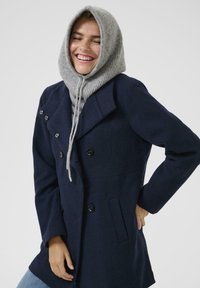 Persona sonriendo que lleva un abrigo azul marino y una capucha de punto gris, de pie con una mano en el bolsillo contra un fondo sencillo.