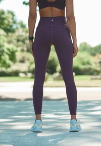 Magas derékú lila leggings rugalmas anyagból, amelyeket karcsú design jellemez, világos színű sportcipővel kiegészítve.