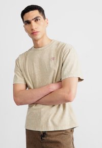 Camiseta de color beige con estampado en tejido suave, cuello redondo, mangas cortas, parche redondo con logo en el pecho, combinada con pantalones marrones texturizados.