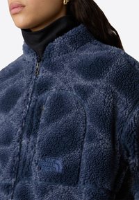 Blå fleecejakke med høy krage, glidelås foran, lap pocket og North Face-logo brodert på brystet.