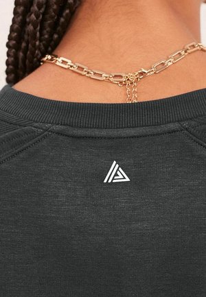 Gros plan sur le cou d'une personne aux cheveux tressés, portant une chaîne en or et une chemise gris foncé arborant un petit logo triangulaire blanc.