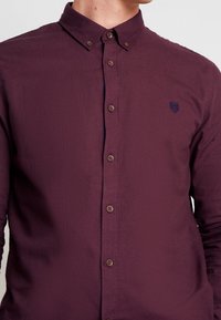 Man met een knoopsluiting donkerpaars overhemd met lange mouwen, een knoop-onder-kraag en een klein geborduurd logo op de borst.