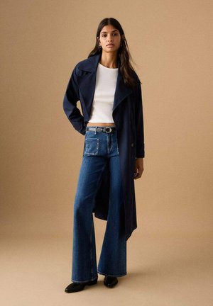 Woman wearing a long navy coat, white cropped ribbed top, blue high-waisted wide-leg jeans with embellished pocket, and black shoes standing.