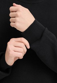 Melnā adītā džemperī ar ribotām piedurknēm. Tuvplānā redzamas rokas, kas pielāgo piedurknes, izceļot tekstūru un piemērotību.