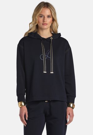 Femme aux longs cheveux blonds portant un sweat à capuche noir avec des cordons dorés, un pantalon noir et des bracelets dorés aux poignets, debout devant un fond uni.