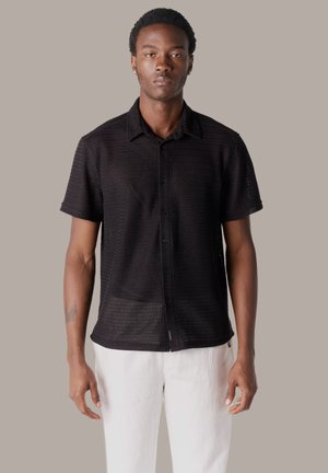 Joven con cabello corto y rizado, vistiendo una camisa negra texturizada de manga corta abotonada y pantalones blancos, de pie con expresión neutral.