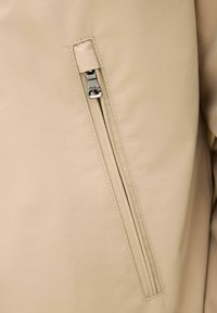 Beige jakke stof med en lodret lynlåslomme, der har en delvist lukket sølvfarvet metal lynlåstrækker.