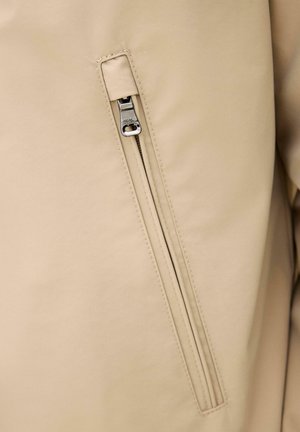 Beige jakke stof med en lodret lynlåslomme, der har en delvist lukket sølvfarvet metal lynlåstrækker.