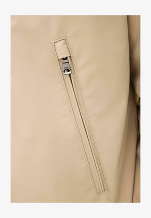 Beige jakke stof med en lodret lynlåslomme, der har en delvist lukket sølvfarvet metal lynlåstrækker.