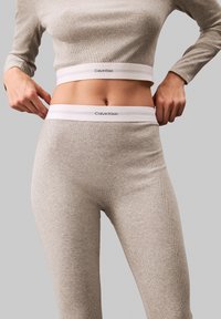 Calvin Klein ribota atpūtas tērps gaiši pelēkā krāsā, ar pieguļošu augšdaļu un legingiem ar baltu zīmola jostu. Gluda tekstūra.