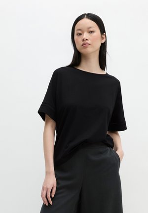 Jeune femme aux longs cheveux noirs, portant un t-shirt noir ample et un pantalon noir, debout avec une main dans la poche devant un fond uni.