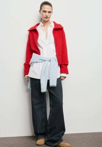Giacca rossa lavorata a maglia con colletto, sopra una camicia bianca con bottoni. Maglione azzurro chiaro annodato in vita, abbinato a jeans scuri a gamba larga.