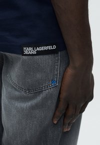 Άτομο με σκουρόχρωμο δέρμα που φορεί γκρι τζιν με μπλε κεντημένο γράμμα "K" στην τσέπη και μπλε πουκάμισο με μαύρο μπάλωμα που γράφει "KARL LAGERFELD JEANS".