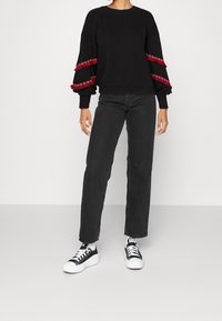 Svart sweatshirt med röda fransar på ärmarna, ribbad textur, rund halsringning, kombinerad med svarta raka jeans och svartvitt sneakers.