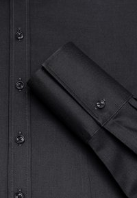 Camicia nera con un tessuto morbido, colletto a punta e chiusura con bottoni neri. I polsini sono piegati con cura.
