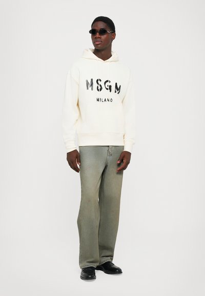 Giovane uomo che indossa occhiali da sole neri, un hoodie crema MSGM Milano, jeans verde sbiadito larghi e scarpe nere, in piedi contro uno sfondo bianco.