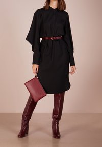 Vestido negro de manga larga con cuello alto y cintura fruncida, combinado con botas de cuero burdeos hasta la rodilla y un clutch a juego.