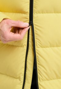 Geel puffer jacket met een zwarte rits. Een hand houdt de rits in de greep terwijl de jas wordt dichtgeritst. Het materiaal lijkt glad en geïsoleerd.