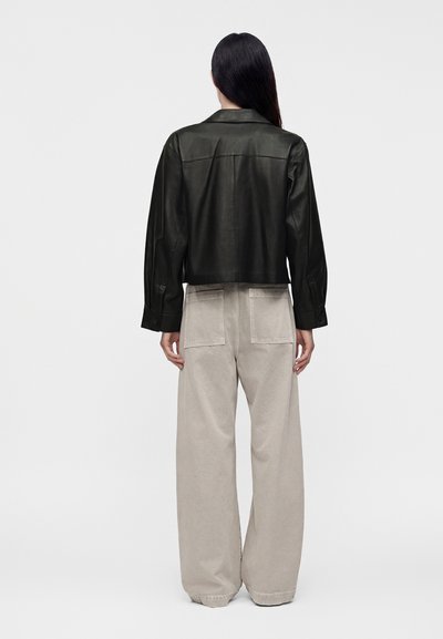 Personne aux longs cheveux foncés porte une veste en cuir noire et un pantalon beige large, debout de dos devant un fond clair uni.