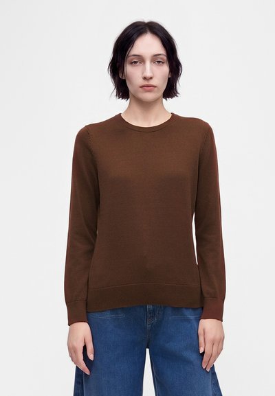 Jeune femme aux cheveux courts et foncés portant un pull marron à manches longues et un jean bleu, debout devant un fond blanc uni.