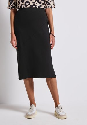 Schwarzer Midirock mit diagonaler Naht, gefertigt aus einem glatten Stoff. Kombiniert mit weißen Turnschuhen für einen lässigen Look.