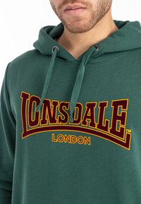 Zeleni dukserica od mekanog materijala s velikim maroon i žutim logotipom "LONSDALE LONDON" preko grudi i podesivom kapuljačom.