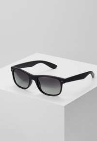 Lunettes de soleil noires avec une finition mate, de forme carrée, verres gris dégradés et logo subtil sur les branches. Texture non réfléchissante.