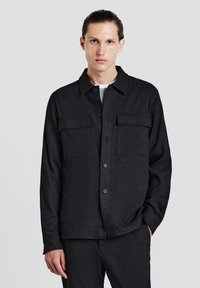 Camicia nera con bottoni realizzata in tessuto testurizzato, dotata di colletto, tasche frontali e una vestibilità comoda. Indossata sopra un top di colore chiaro.