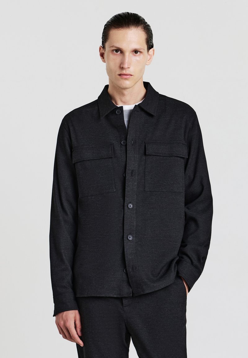 Camicia nera con bottoni realizzata in tessuto testurizzato, dotata di colletto, tasche frontali e una vestibilità comoda. Indossata sopra un top di colore chiaro.