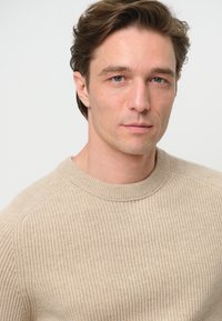 Beige ribbad tröja med rund halsringning, med en texturerad stickdesign, bärs av en modell mot en neutral bakgrund.