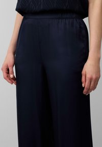 Pantalons en satin bleu marine avec une taille élastique, présentant une texture lisse et un design à jambes larges. Main reposant légèrement sur le tissu.