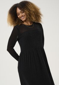 Black long-sleeve dress with a sheer top section, featuring a pleated skirt and a subtle shimmer. Accented with a delicate necklace.