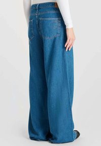 Pantalones de mezclilla de pierna ancha en azul con cintura alta, que cuentan con dos bolsillos traseros y un sutil detalle de costura. Textura suave, diseño casual.