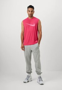 Miesten hihaton vaaleanpunainen Nike-treenitoppi, jossa on swoosh-kuvio, yhdistetty vaaleanharmaisiin collegehousuihin ja valkoisiin lenkkareihin.