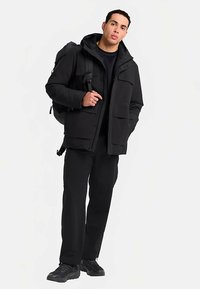 Schwarze gepolsterte Jacke mit Kapuze, zwei Fronttaschen und durchgehendem Reißverschluss. Kombiniert mit schwarzer Hose und einem Rucksack. Glatte Stoffstruktur.