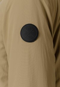 Tessuto beige con una texture liscia. Presenta una patch rotonda in pelle nera con il simbolo in rilievo "MT 97" sulla parte superiore del braccio.