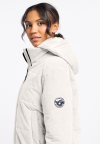 Lichtgrijze corduroy pufferjack met een capuchon, met een gequilt ontwerp en een rond logo accent op de arm. Voorzien van een ritssluiting aan de voorkant.