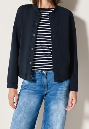 Frau trägt eine marineblaue Jacke mit Knöpfen über einem schwarz-weiß gestreiften Hemd und hellblauen Jeans.