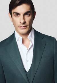 Ciemnozielona marynarka o fakturowanej powierzchni, noszona na białej koszuli. Charakterystyczny dekolt w serek i klapy z nacięciem. Portret zbliżeniowy.