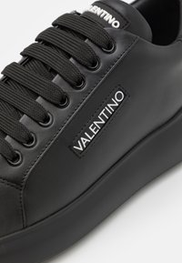 Black leather sneaker featuring flat black laces, a thick sole, and a white "VALENTINO" logo tag stitched on the side.