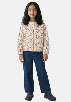 Fille aux cheveux noirs mi-longs portant une veste blanche à motifs de cerises, un jean bleu avec des dessins de cœurs rouges, et des bottes noires, debout face à l'avant.